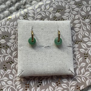May include: A pair of gold hoop earrings with round, light green jade pendants. The earrings are displayed on a small, square, off-white linen display card. The background features a gray and white floral pattern.