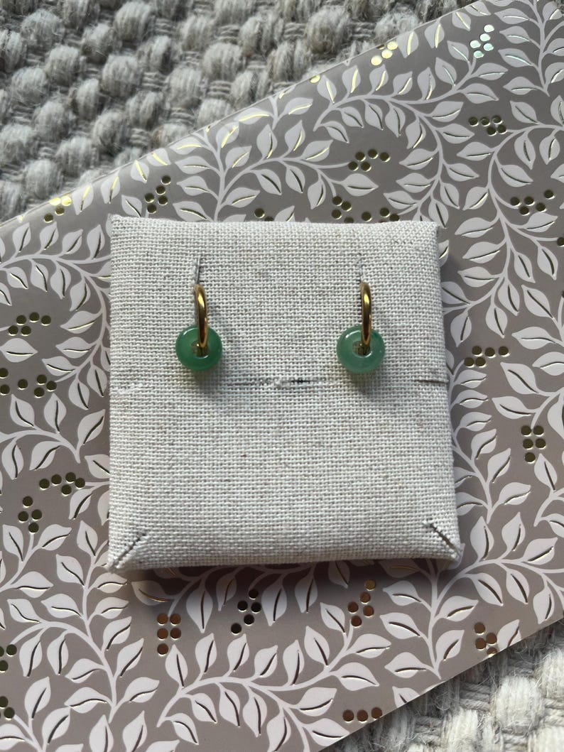 May include: A pair of gold hoop earrings with round, light green jade pendants. The earrings are displayed on a small, square, off-white linen display card. The background features a gray and white floral pattern.