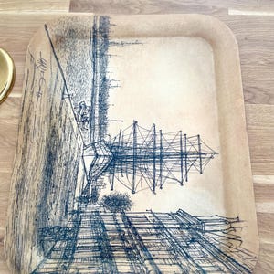 May include: A vintage tray with a black and white illustration of a ship docked in a city. The tray is made of a light brown material and has a slightly raised edge.