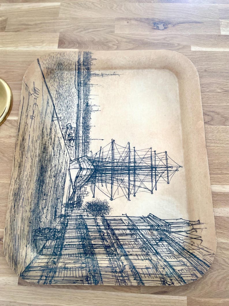 May include: A vintage tray with a black and white illustration of a ship docked in a city. The tray is made of a light brown material and has a slightly raised edge.