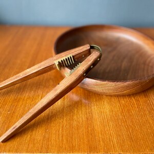 May include: A wooden nutcracker with a brass mechanism, resting on a wooden bowl. The nutcracker has a long, thin handle and a small, round head with a serrated edge.