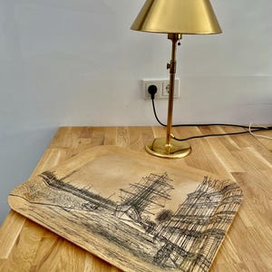 May include: A wooden tray with a black and white illustration of a ship and buildings. The tray is sitting on a wooden table next to a gold table lamp.