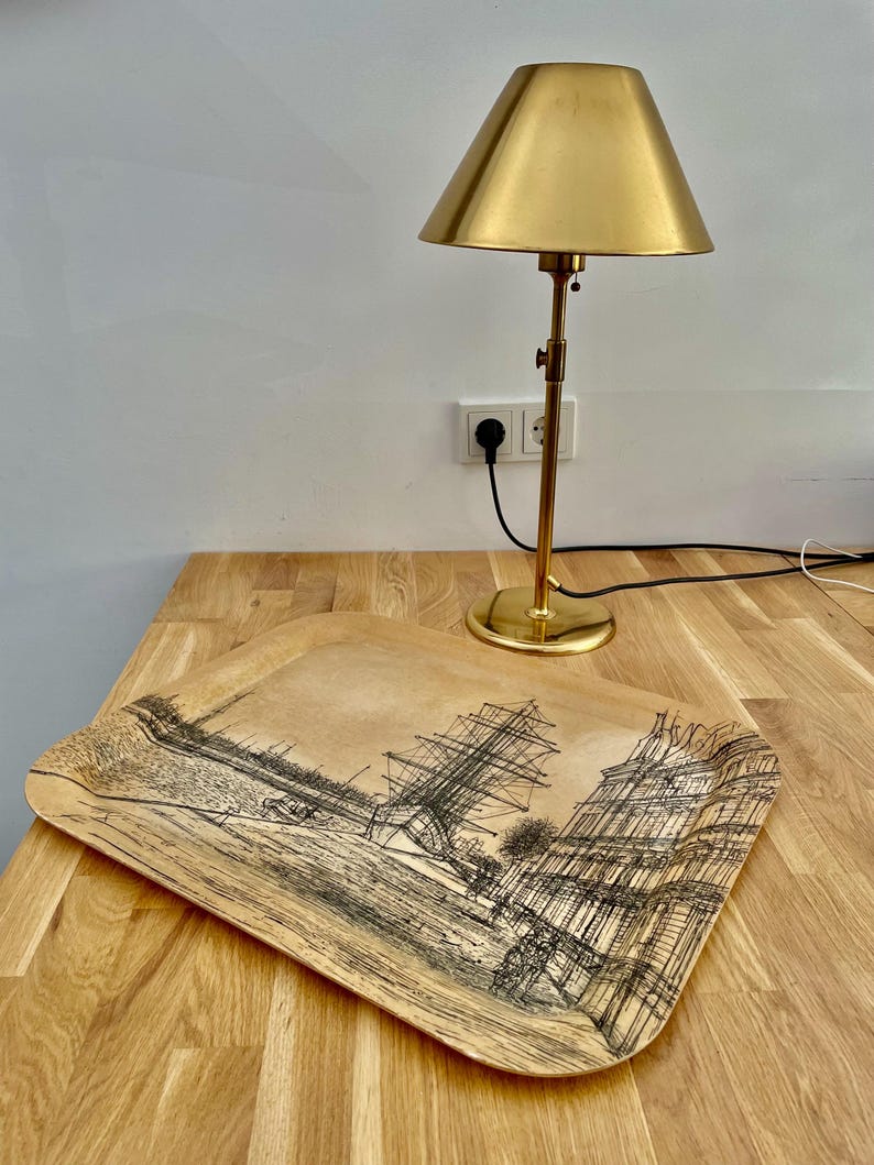 May include: A wooden tray with a black and white illustration of a ship and buildings. The tray is sitting on a wooden table next to a gold table lamp.
