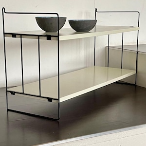 Puede incluir: Estantería de dos niveles con estructura de metal negro y estantes blanquecinos. Dos cuencos grises oscuros con interiores azules se encuentran en el estante superior. Ideal para almacenamiento o exhibición.