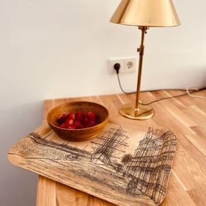 May include: A wooden tray with a black and white illustration of a ship and buildings. A small wooden bowl filled with red strawberries sits on the tray. A gold table lamp with a gold shade is in the background.