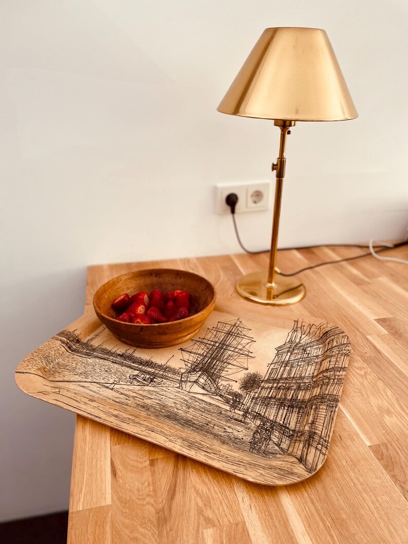 May include: A wooden tray with a black and white illustration of a ship and buildings. A small wooden bowl filled with red strawberries sits on the tray. A gold table lamp with a gold shade is in the background.