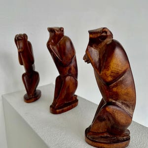 May include: Three wooden monkey sculptures in a row, each in a different pose. The monkeys are carved from a dark brown wood and are on a white surface. The sculptures represent the "see no evil, hear no evil, speak no evil" concept.