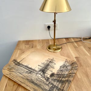 May include: A wooden tray with a black and white illustration of a ship and buildings. The tray is sitting on a wooden table next to a gold table lamp.