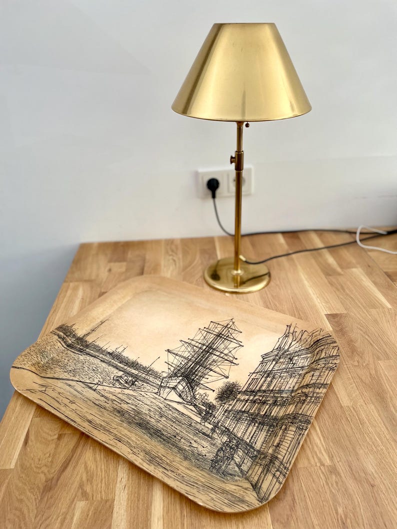 May include: A wooden tray with a black and white illustration of a ship and buildings. The tray is sitting on a wooden table next to a gold table lamp.