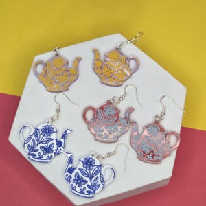 Könnte beinhalten: Sechs Paar ohrringförmige Teekannen in verschiedenen Farben. Die Ohrringe haben Blumendesigns in Gelb, Rosa, Blau und Weiß. Die Ohrringe werden auf einer weißen geometrischen Oberfläche vor einem gelben und rosa Hintergrund präsentiert.