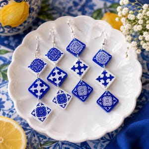 Puede incluir: Cuatro pendientes colgantes con una serie de azulejos en forma de diamante conectados en azul y blanco. Cada pendiente consta de tres azulejos apilados con intrincados patrones florales y de cruz, suspendidos de ganchos de metal plateado.
