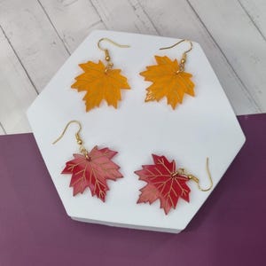 May include: Four pairs of leaf-shaped earrings. Two pairs are yellow and two are red, all with gold-coloured hooks. The earrings are displayed on a white hexagonal surface against a light wood-grain background.