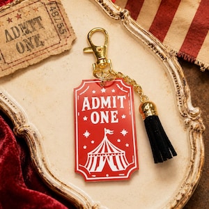 Könnte beinhalten: Ein roter Acryl-Schlüsselanhänger in Ticketform mit weißem Text ADMIT ONE und einer Zirkuszelt-Grafik. Der Schlüsselanhänger verfügt über einen goldfarbenen Karabinerverschluss, eine kurze Kette und eine schwarze Stoffquaste an der Seite.