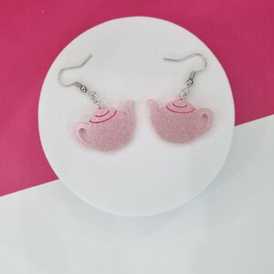 Puede incluir: Un par de pendientes de tetera rosa con purpurina y ganchos plateados. Las teteras tienen un contorno rojo.