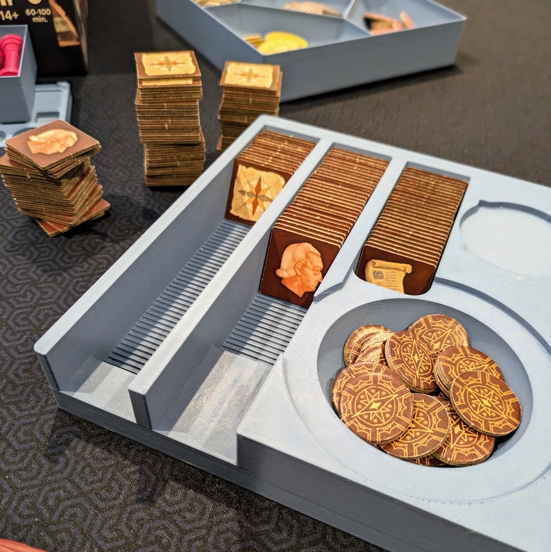 Peut inclure: Un plateau organisateur de jeu de soci&eacute;t&eacute; en plastique bleu clair avec plusieurs compartiments. Le plateau contient des piles de tuiles carr&eacute;es en carton illustr&eacute;es de boussoles, de profils humains et de parchemins, ainsi qu'une section circulaire remplie de jetons ronds.
