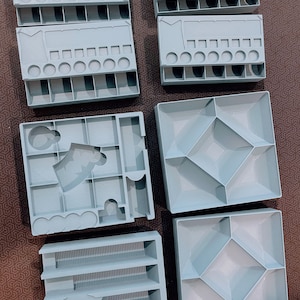 Peut inclure: Un ensemble de sept organisateurs de jeux de soci&eacute;t&eacute; en plastique gris clair, comprenant divers compartiments, des fentes rectangulaires, des indentations circulaires et des sections carr&eacute;es pour trier les jetons et &eacute;l&eacute;ments de jeu.