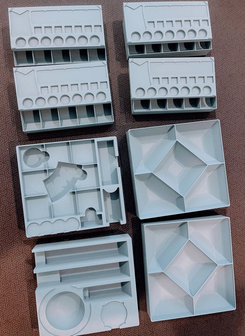 Peut inclure: Un ensemble de sept organisateurs de jeux de soci&eacute;t&eacute; en plastique gris clair, comprenant divers compartiments, des fentes rectangulaires, des indentations circulaires et des sections carr&eacute;es pour trier les jetons et &eacute;l&eacute;ments de jeu.