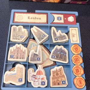 Peut inclure: Un plateau organisateur de jeu de soci&eacute;t&eacute; en plastique bleu contenant divers composants en carton. Le plateau comporte des compartiments avec des jetons de b&acirc;timents illustr&eacute;s num&eacute;rot&eacute;s, des jetons orange circulaires avec des chiffres romains et une tuile rectangulaire marqu&eacute;e London.