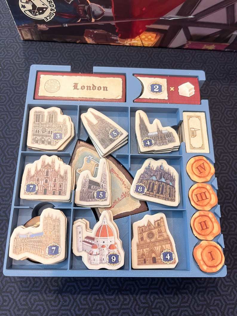 Peut inclure: Un plateau organisateur de jeu de soci&eacute;t&eacute; en plastique bleu contenant divers composants en carton. Le plateau comporte des compartiments avec des jetons de b&acirc;timents illustr&eacute;s num&eacute;rot&eacute;s, des jetons orange circulaires avec des chiffres romains et une tuile rectangulaire marqu&eacute;e London.