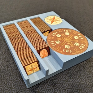 Peut inclure: Un plateau organisateur de jeu de soci&eacute;t&eacute; en plastique bleu clair contenant trois fentes verticales remplies de tuiles en bois carr&eacute;es. Un cadran circulaire en carton marron avec des motifs de d&eacute;s blancs et les chiffres 1 &agrave; 6 est plac&eacute; &agrave; droite.