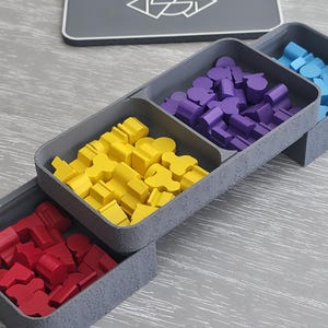 Puede incluir: Un conjunto de bandejas de almacenamiento de plástico gris para componentes de juegos de mesa, con tres compartimentos conectados llenos de meeples de madera rojos, amarillos y morados, y una tapa gris con un logo geométrico blanco.