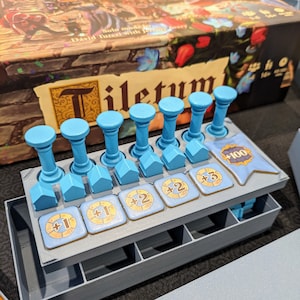 Peut inclure: Un plateau organisateur de jeu de soci&eacute;t&eacute; contenant six piliers cylindriques bleu clair, cinq jetons en forme de maison bleu clair et cinq tuiles carr&eacute;es avec des valeurs num&eacute;riques plus un, plus deux et plus trois, ainsi qu'un jeton drapeau bleu marqu&eacute; plus 100.