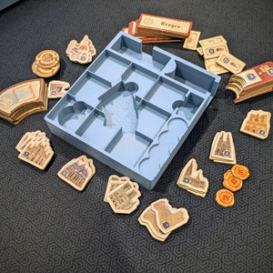 Peut inclure: Un plateau organisateur de jeu de soci&eacute;t&eacute; en plastique bleu clair avec plusieurs compartiments, entour&eacute; de divers jetons de jeu en carton, de cartes marqu&eacute;es Troyes et de petits marqueurs circulaires avec des chiffres romains.