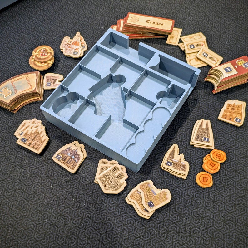 Peut inclure: Un plateau organisateur de jeu de soci&eacute;t&eacute; en plastique bleu clair avec plusieurs compartiments, entour&eacute; de divers jetons de jeu en carton, de cartes marqu&eacute;es Troyes et de petits marqueurs circulaires avec des chiffres romains.