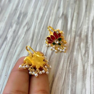 May include: Gold-colored earrings with a lotus flower design. The earrings feature red and green enamel detailing, small white pearls, and a hook closure. The earrings are displayed on a textured, light gray background.