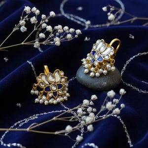 May include: A pair of gold-colored earrings with a lotus flower design, featuring white and blue enamel and small pearl accents. The earrings are displayed on a dark blue velvet surface with delicate white flowers.