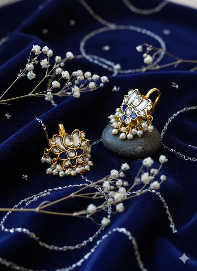 May include: A pair of gold-colored earrings with a lotus flower design, featuring white and blue enamel and small pearl accents. The earrings are displayed on a dark blue velvet surface with delicate white flowers.
