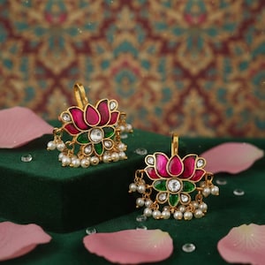 May include: A pair of gold-coloured lotus flower earrings. Each earring features pink and green enamel petals, a central clear stone, and dangling faux pearls. The earrings are on a dark green surface.