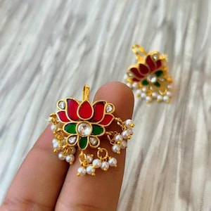 May include: Gold-toned lotus flower earrings with red, green, and white enamel detailing. The earrings feature a central clear stone and dangling faux pearls. The design is intricate and ornate, with a focus on floral elements.