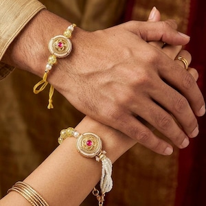 Könnte beinhalten: Zwei dekorative Armbänder mit einem zentralen goldfarbenen runden Medaillon mit einem rosa Steineinsatz. Ein Armband hat eine gelbe Kordel mit gelben Perlen, das andere weiße perlenartige Kügelchen und einen hängenden goldfarbenen Glöckchenanhänger.