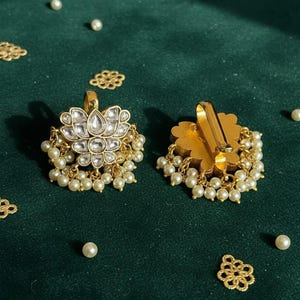 May include: Gold-toned earrings with a floral design, adorned with clear gemstones and dangling faux pearls. The earrings are set against a dark green backdrop, with scattered pearls and decorative gold accents.