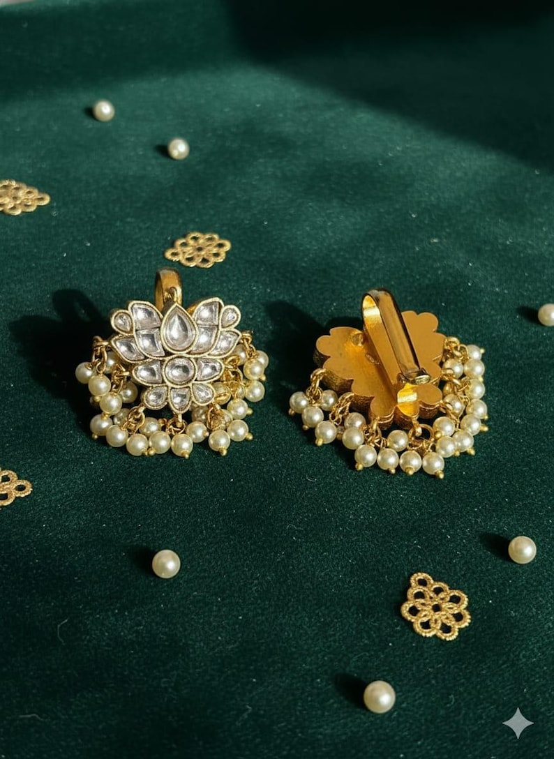 May include: Gold-toned earrings with a floral design, adorned with clear gemstones and dangling faux pearls. The earrings are set against a dark green backdrop, with scattered pearls and decorative gold accents.