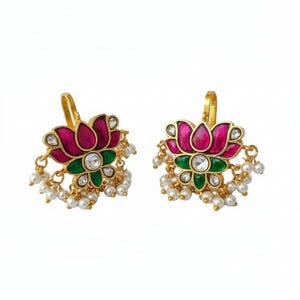 May include: A pair of gold-colored earrings featuring a lotus flower design. The earrings have a magenta-colored flower with green accents and a central clear stone. Small white pearls dangle from the bottom of each earring.