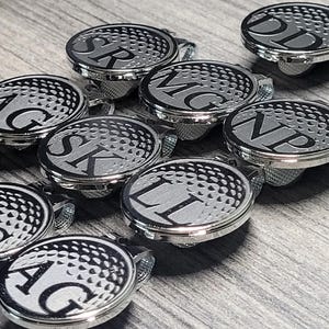 May include: A set of nine silver golf ball hat clips with black engraved initials. Each clip features a textured golf ball design.