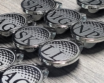 Puede incluir: Un conjunto de nueve clips para sombreros de bola de golf plateados con iniciales grabadas en negro. Cada clip presenta un diseño de bola de golf texturizado.