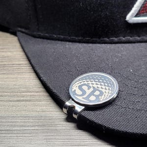 Peut inclure: Un clip de chapeau en forme de balle de golf argenté avec les initiales "SB" gravées dessus. Le clip est fixé à une casquette de baseball noire.