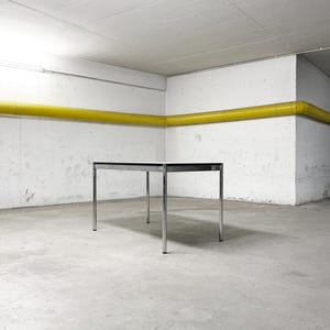 Könnte beinhalten: Ein rechteckiger Tisch mit einer glatten weißen Tischplatte und einem polierten Chrom-Metallrahmen mit vier schlanken Beinen. Der Tisch hat ein schlichtes Design mit einer schwarzen Kante um die weiße Tischplatte, was für ein neutrales Aussehen sorgt.