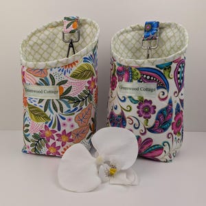 Könnte beinhalten: Zwei Wandorganizer aus Stoff mit floralen Mustern in Rosa, Blau und Grün. Jeder hat einen Metallhaken und ein Etikett mit der Aufschrift "Greenwood Cottage". Die Organizer stehen auf einer weißen Oberfläche mit weißen Orchideenblüten.