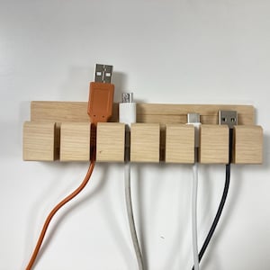 Könnte beinhalten: Ein Kabel-Organizer aus Holz mit fünf Schlitzen, der verschiedene USB-Kabel in Orange, Weiß und Schwarz hält. Der Organizer besteht aus hellem Holz und ist auf einer weißen Oberfläche montiert.