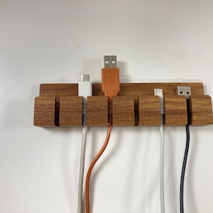 Könnte beinhalten: Ein hölzerner Kabelorganizer mit sechs Steckplätzen zum Halten von Kabeln. Der Organizer ist an einer weißen Wand montiert. Vier Kabel sind in den Organizer gesteckt, zwei sind weiß, eines ist orange und eines ist schwarz.
