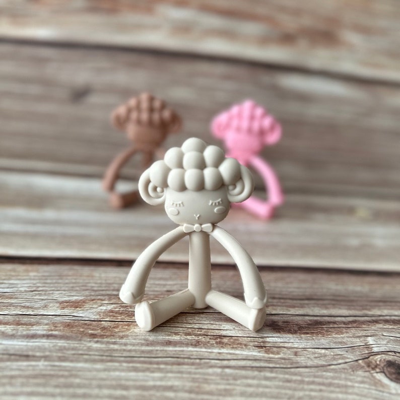 May include: A beige silicone baby teether shaped like a sheep with a bow tie. The teether has a textured surface and is designed for babies to chew on.