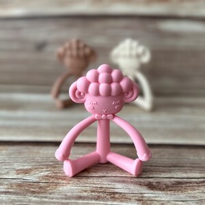 May include: A pink silicone baby teether shaped like a sheep with a bow tie. The teether has a textured head and long arms and legs.