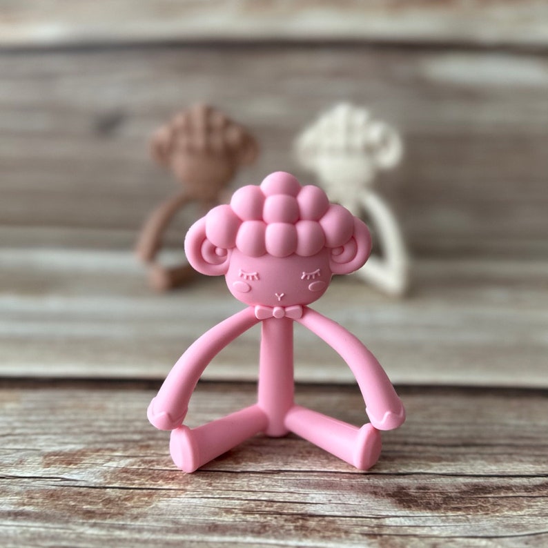 May include: A pink silicone baby teether shaped like a sheep with a bow tie. The teether has a textured head and long arms and legs.