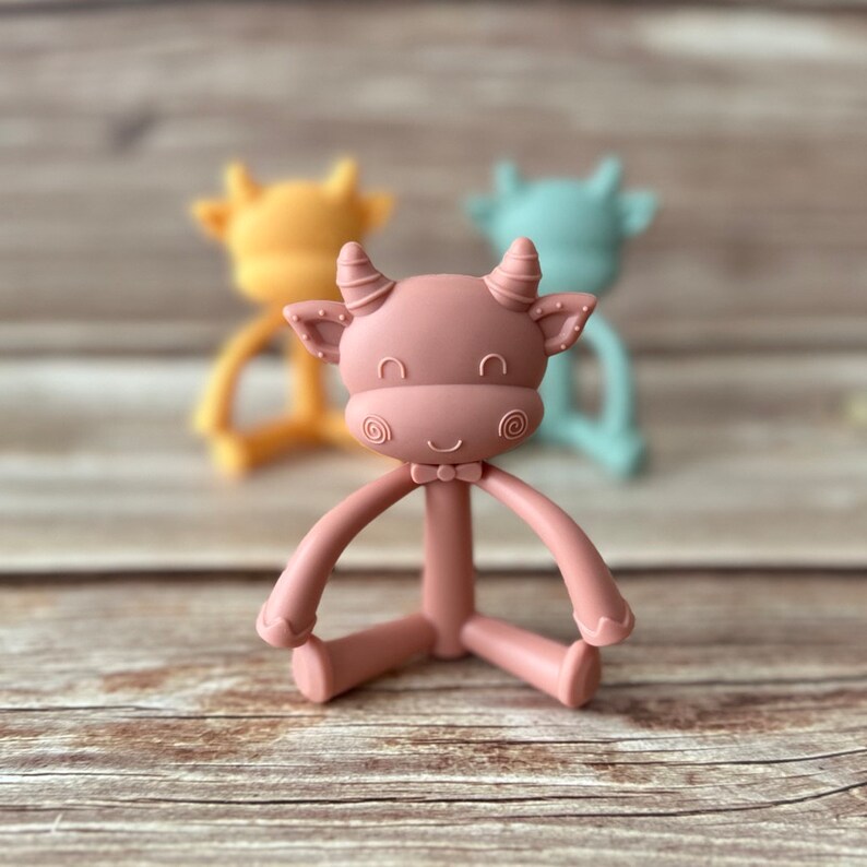 May include: A pink silicone baby teether shaped like a cow with a bow tie. The teether has a long neck and four arms that can be bent and twisted.