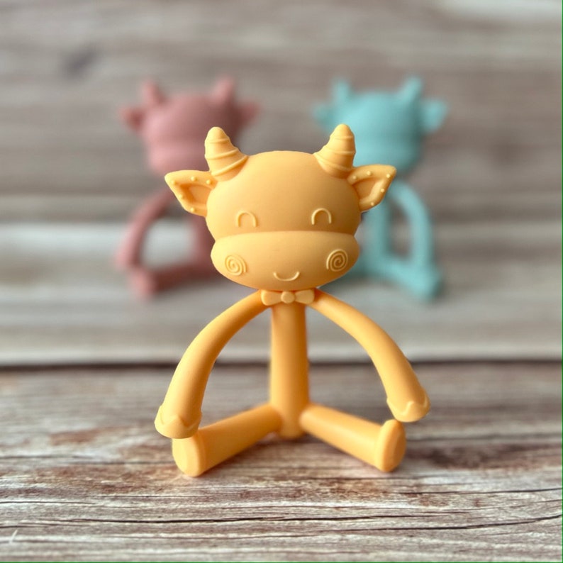May include: A yellow silicone baby teether shaped like a cow with a bow tie. The teether has flexible arms and legs that can be twisted and turned.