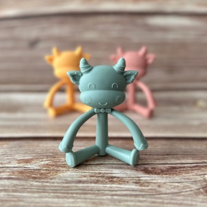 May include: A teal blue silicone baby teether shaped like a cow with a bow tie. The teether has a textured surface and is designed for babies to chew on.